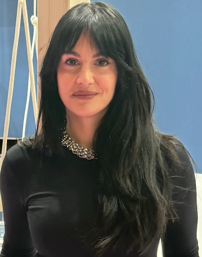 Professional portrait of a woman with long dark hair wearing a black top and decorative necklace against a blue background