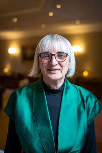 Professional portrait of a woman with white hair and glasses wearing a teal cardigan over a black turtleneck, smiling in a warmly lit interior setting.