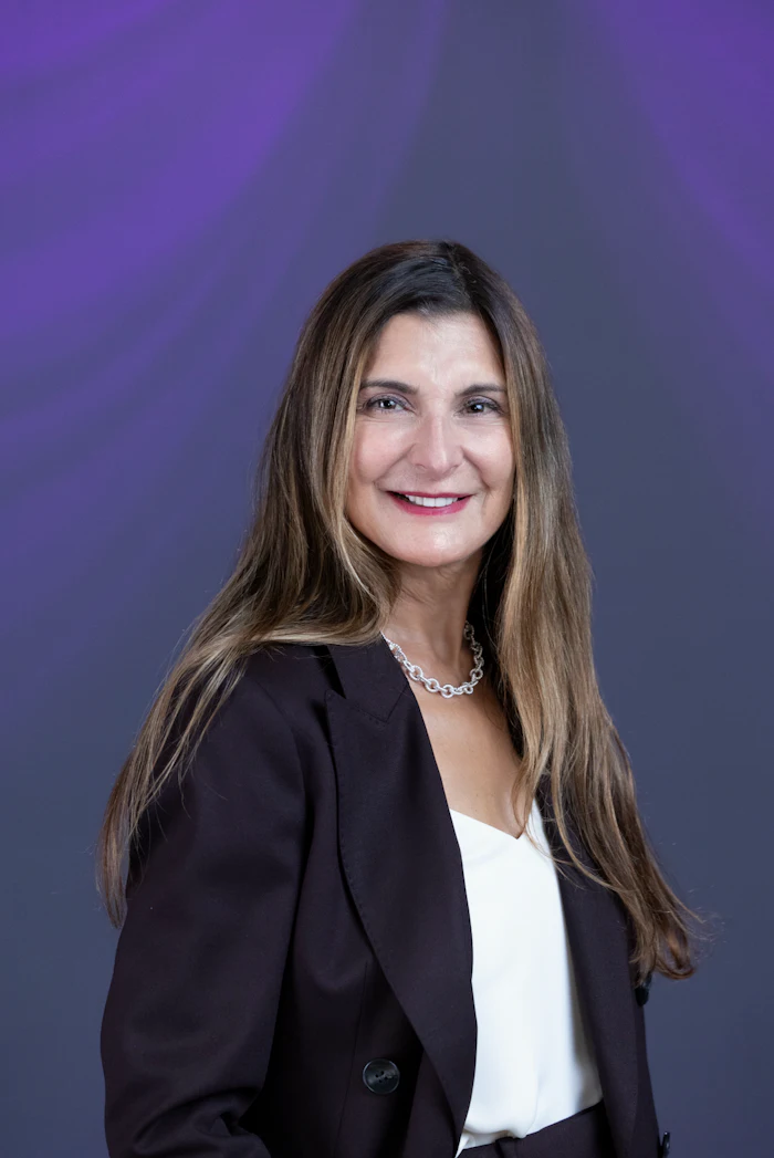 Professional headshot of a woman in black blazer and white blouse against purple background