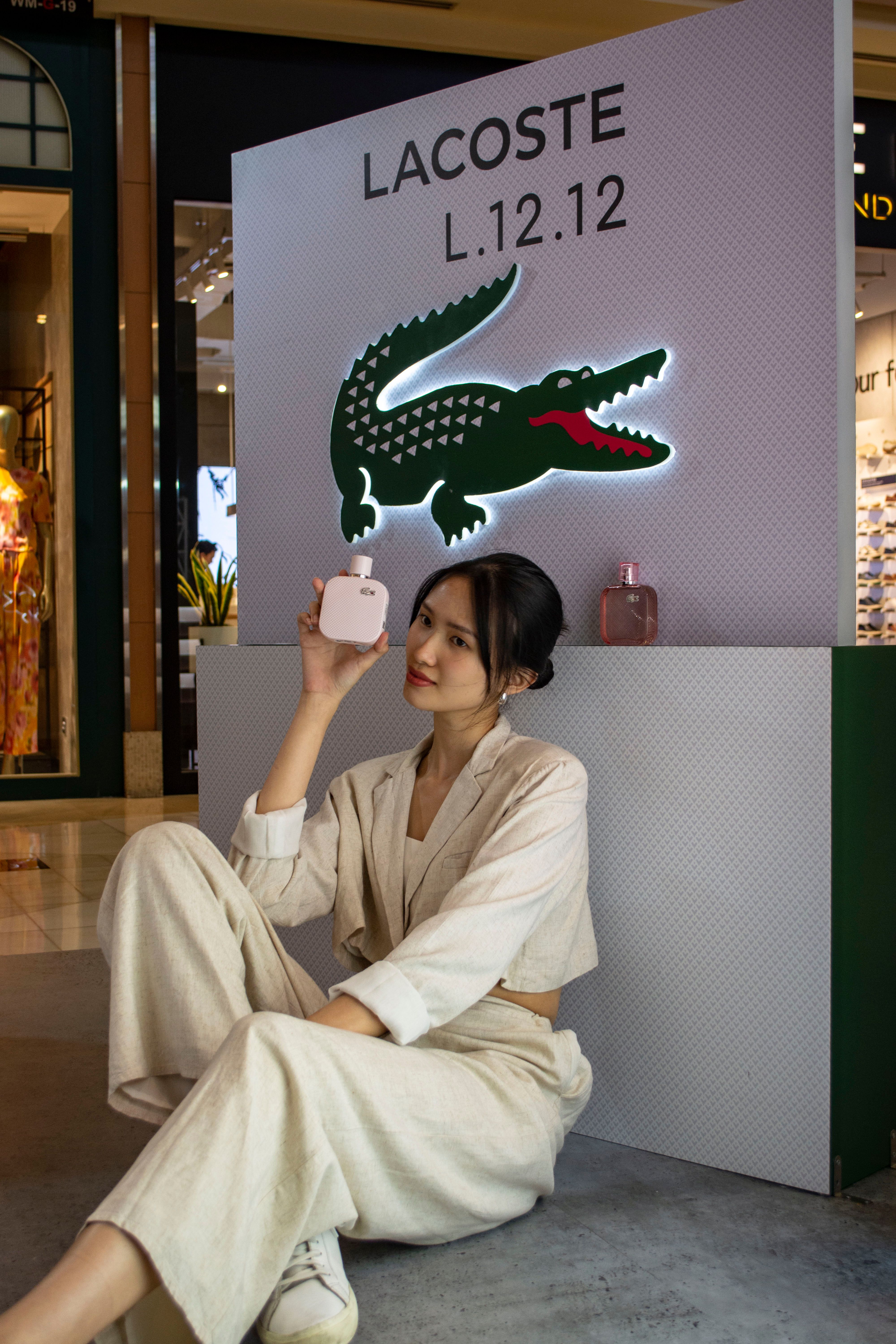 Woman in cream suit holding Lacoste fragrance bottle at L.12.12 promotional display with iconic green crocodile logo