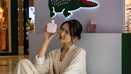 Woman in cream suit holding Lacoste fragrance bottle at L.12.12 promotional display with iconic green crocodile logo