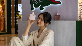 Woman in cream suit holding Lacoste fragrance bottle at L.12.12 promotional display with iconic green crocodile logo