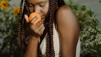 Woman with braided hair eating an orange surrounded by green plants and yellow flowers