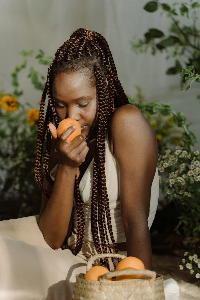 Woman with braided hair eating an orange surrounded by green plants and yellow flowers