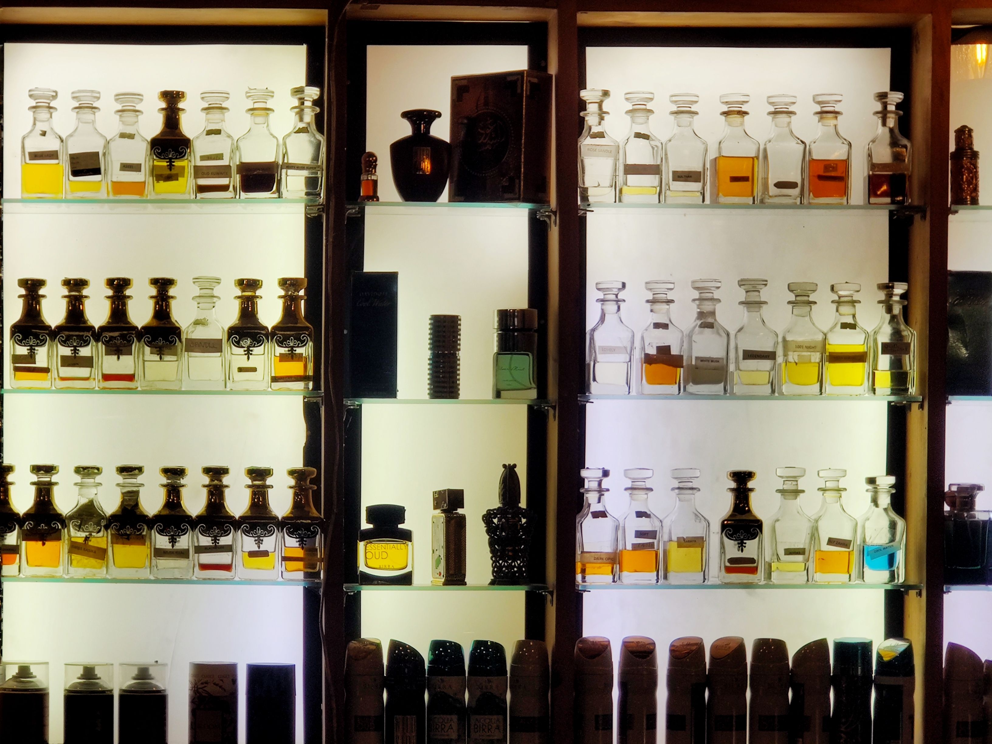 Illuminated wooden cabinet displaying collection of perfume and fragrance bottles in various sizes and colors arranged on multiple shelves