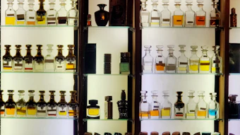 Illuminated wooden cabinet displaying collection of perfume and fragrance bottles in various sizes and colors arranged on multiple shelves