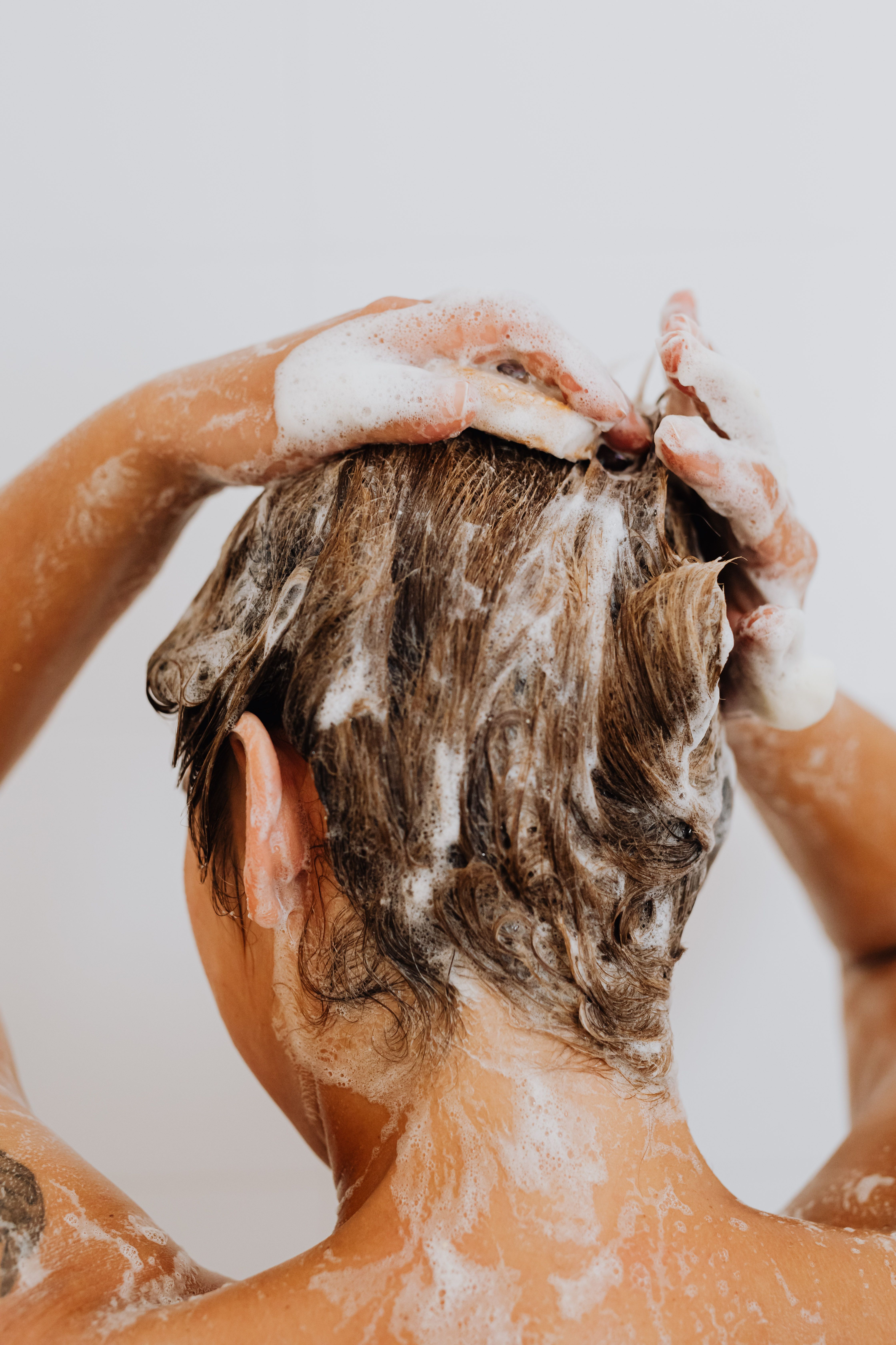 Person lathering hair with shampoo while washing, hands visible applying white foam to wet brown hair