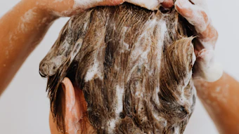 Person lathering hair with shampoo while washing, hands visible applying white foam to wet brown hair