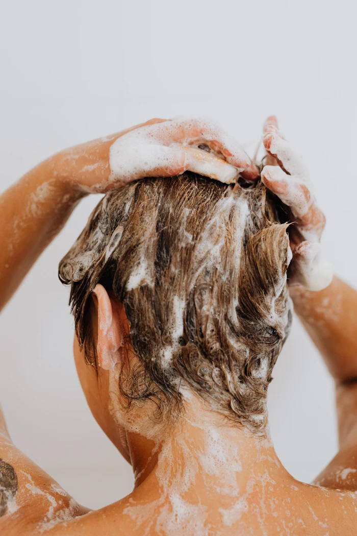 Person lathering hair with shampoo while washing, hands visible applying white foam to wet brown hair