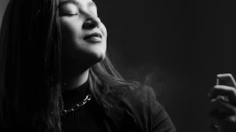 Black and white portrait of a woman with long dark hair, eyes closed, wearing a dark textured jacket and chain necklace against a dark background