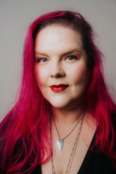 Professional portrait of a woman with vibrant magenta hair, red lipstick, and layered silver necklaces against a neutral background