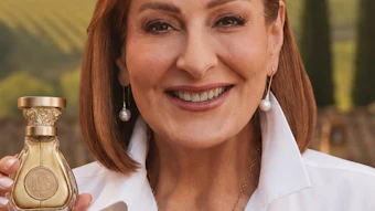 Woman with auburn hair holding a gold Baccarat fragrance bottle, smiling at camera against vineyard background