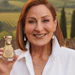 Woman with auburn hair holding a gold Baccarat fragrance bottle, smiling at camera against vineyard background